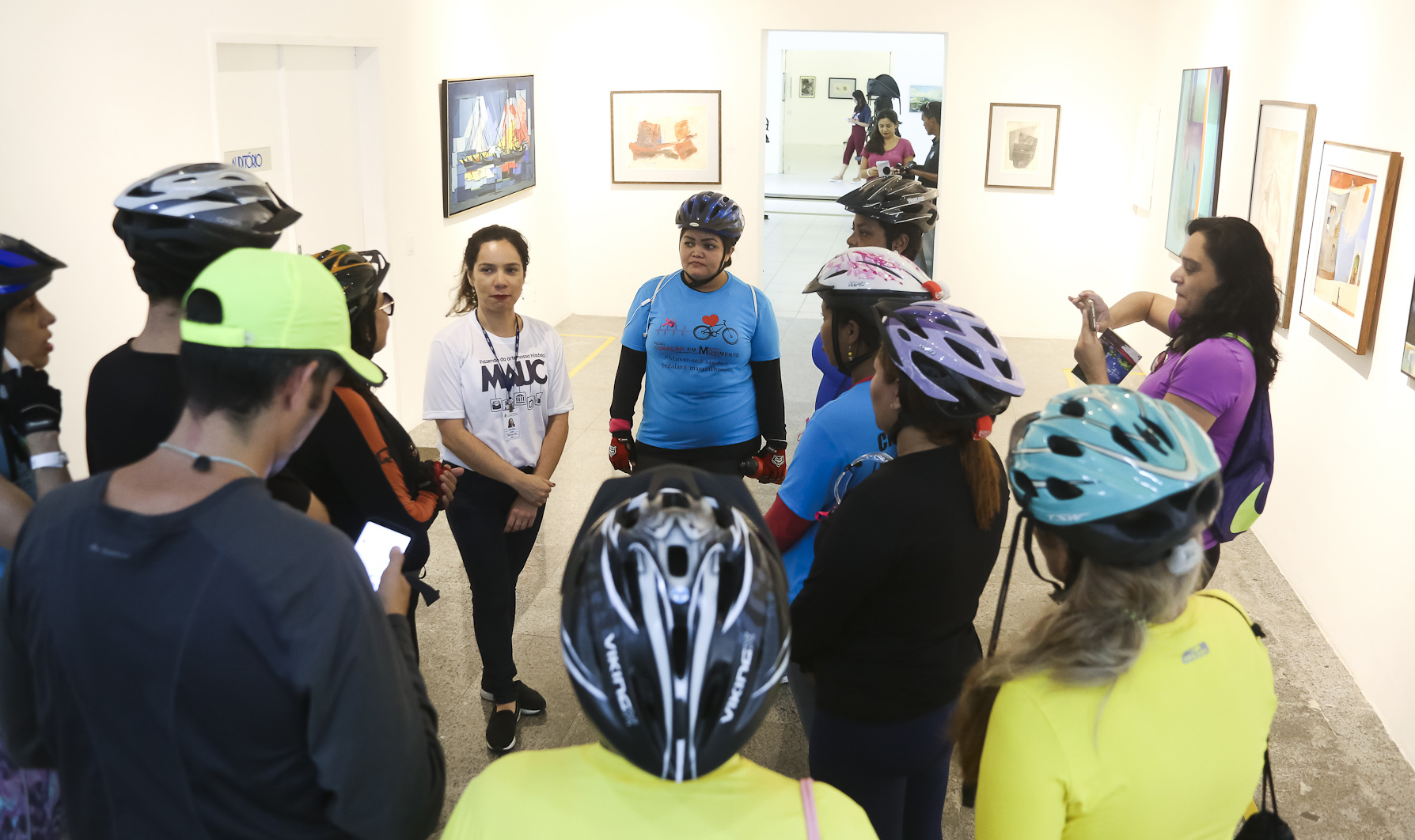 Grupo de ciclistas paramentados com capacetes conversam dispostos em círculo junto a uma monitora do Museu de Arte da UFC